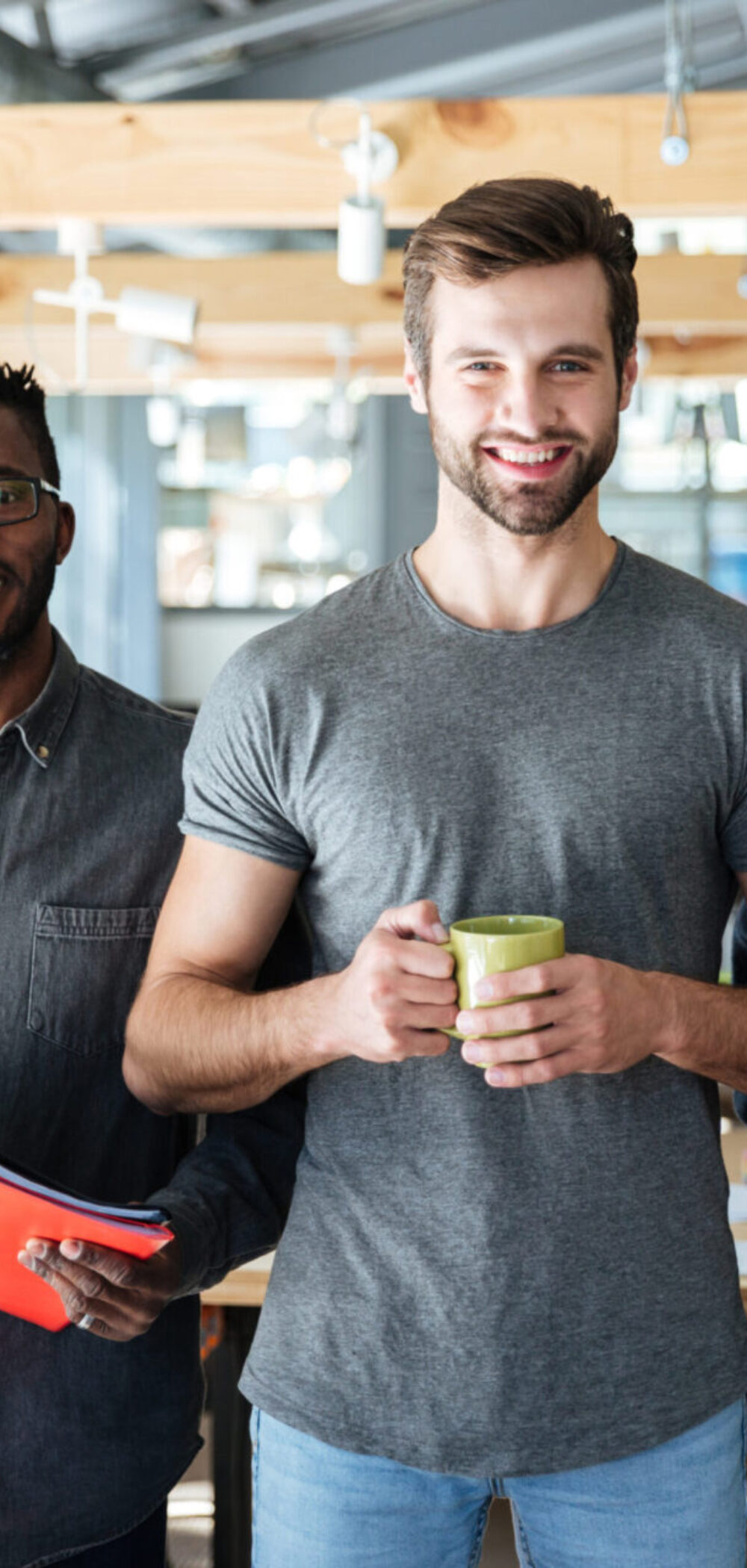 Photo of happy young colleagues in office coworking standing holding laptop, documents and coffee. Looking at camera.